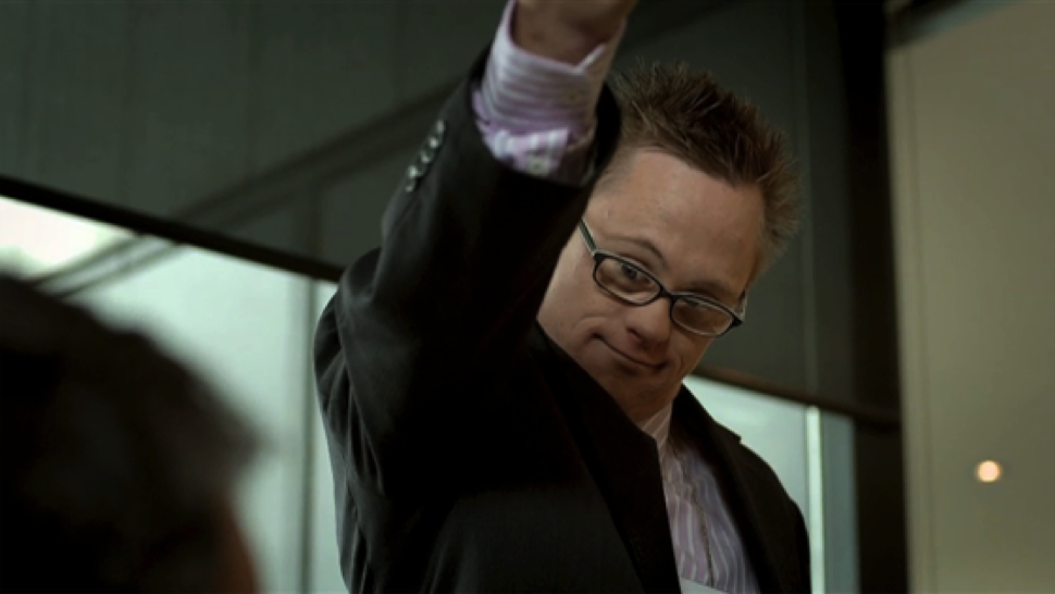 A caucasian man with Down syndrome wearing glasses and a business suit contently smiles at the camera as he pumps his fist in his air.